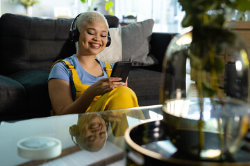 Happy fashionable unaltered biracial woman sitting on floor at home in headphones using smartphone