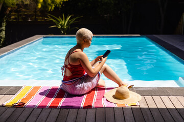 Happy fashionable unaltered biracial woman sitting by swimming pool using smartphone in sunny garden