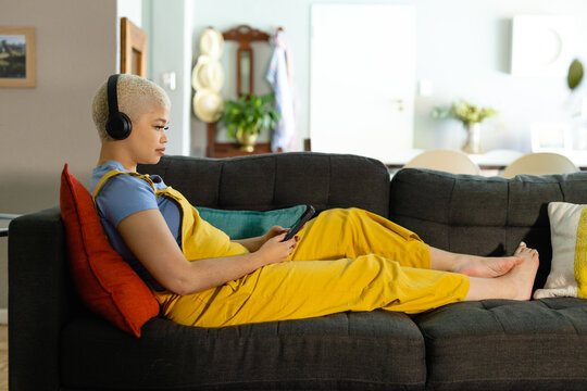 Fashionable unaltered biracial woman relaxing on couch at home in headphones using smartphone