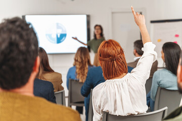 During a SWOT analysis presentation in a large office room, a red-haired businesswoman raises her hand to ask a question. The diverse audience is viewed from the back.
