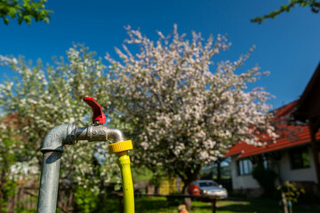 Garden water tap with a yellow hose at a residential home at springtime, flowering fruit trees in...