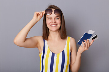 Naklejka premium Smiling cheerful woman in swimsuit holding passport isolated on gray background, booking tickets online, raised her sunglasses, joyful expression.