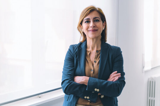 Portrait of a Caucasian senior businesswoman in her 50s. She is dressed in casual office attire and standing near a window in a white office room. With her arms crossed, she is smiling at the camera.