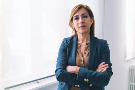 Portrait of a Caucasian senior businesswoman. She is dressed in casual office attire and standing near a window in a white office room. With her arms crossed, she is looking seriously at the camera.