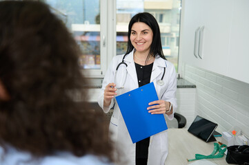 Confident portrait of a Caucasian charming pretty woman, female doctor, GP, physician, gynecologist in white medical gown, smiling broadly, welcoming her patient at appointment in new medical clinic