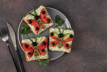 Vegetarian sandwiches form butterfly for kids with tomato, avocado and olives on gray plate on brown table, Top view