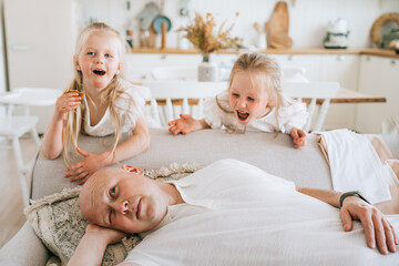 Bald caucasian man laying on couch tries to relax, sleep while little daughters misbehaving screaming at living room. Naughty little blonde sisters mischievous at home. Childhood, tired father.