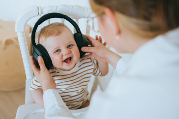 Sister entertains little brother puts on his head headphones. Little child boy enjoying music sitting in children chair. Newborn baby smiles likes listening music. Childhood.
