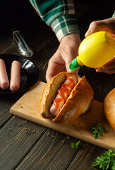 Cooking hot dogs by the hands of a cook on the kitchen table. The concept of cooking fast food or street food. The chef adds mustard for flavor