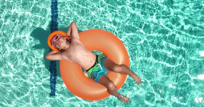 Top view of happy young child enjoying summer vacation outdoors in water in the swimming pool. Cute little kid in swimming suit and sunglasses relaxing on an inflatable ring. Kid floating in a pool.