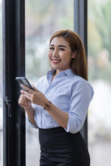 Fototapeta premium Smiling business Asian woman using her phone in the office. Small business entrepreneur looking at her mobile phone and smiling while communicating with her office colleagues