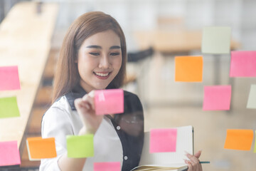 Young Asian woman Creative team use post it notes to share idea sticky note on glass wall. Asian business people design planning and Brainstorming thinking sticky History notes concept.
