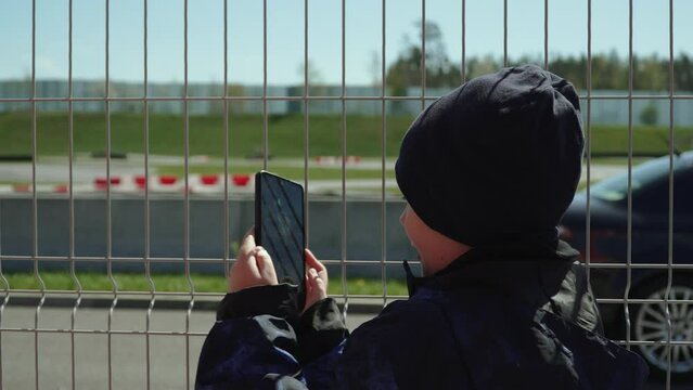 Boy is trying to shoot video on smartphone of car passing through gaps in metal fence along race track. On cool spring day, child uses their wits to film blog post.
