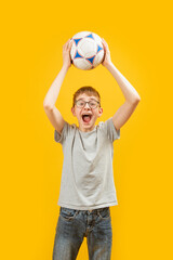 Cheerful boy in jeans and T-shirt holds soccer ball above his head. Teenager is football fan. Schoolboy with ball glasses.