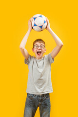Cheerful boy in jeans and T-shirt holds soccer ball above his head. Teenager is football fan....