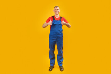 Fototapeta na wymiar Cheerful builder in blue jumpsuit shows thumbs up and smiles. Full-length portrait of worker showing like gesture. Copy space.