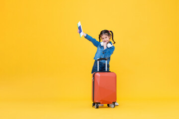 Cute little girl passenger with baggage holding passport and boarding pass in isolated yellow color background studio shot, minor travel concept