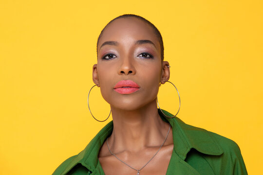 Headshot portrait of elegant fashionable skinhead African American woman face with big earrings against yellow color background studio shot