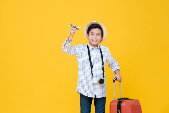 Young smiling Asian boy with baggage and camera in isolated yellow background studio shot, minor travel concept