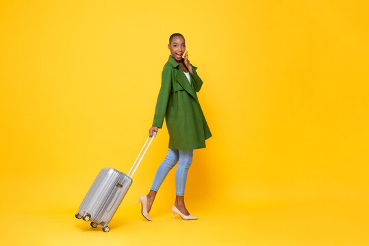 Fashionable skinhead African American woman traveler walking with trolley bag ready to fly against yellow color isolated background studio shot