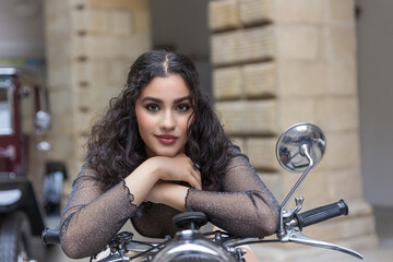 Tanned young woman with long black hair is posing on the vintage motorcycle leaning on the handlebars.  Horizontally. 