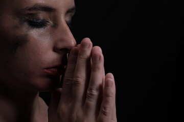 Fototapeta na wymiar Portrait of a young crying woman with smeared mascara and lipstick praying on a black background
