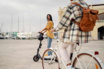 A happy hipster woman on an electric scooter is looking at her friend on a bike and having fun...