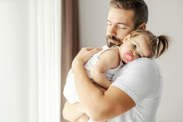 Portrait of a father holding his baby girl in his arms and putting her to sleep. Baby is holding her head on dad's shoulder and looking at the camera.