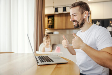 A happy casual businessman is giving thumbs up for project while having online briefing while babysitting his infant.