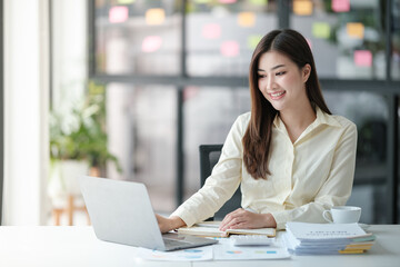 Photo of attractive woman sitting at desk working on computer looking at screen, smiling, wearing yellow shirt and glasses, typing on keyboard, holding a cup of coffee.