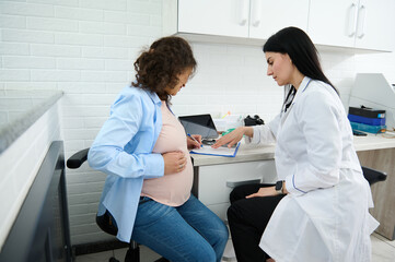 Young pregnant woman putting hand on her belly, sitting in a doctor's office and signing a contract...