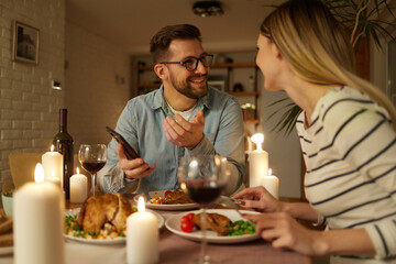 Happy couple enjoying food and wine while surfing the net on smart phone at home