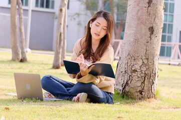 Happy smiling young woman sitting and studying on laptop under tree.