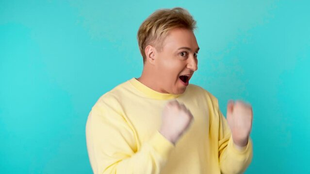 Face, winner celebration of man, fist pump gesture. Emotional guy male excited after success, winning, victory or news, showing Yes expression in yellow sweatshirt, studio isolated on blue background