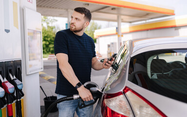 Handsome bearded man refueling car and looking on the scoreboard while standing on self service gas...