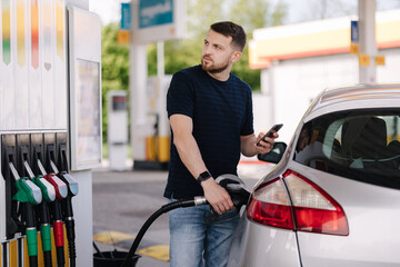 Bearded man refuelling car on gas station and looking into his smartphone. Man compares fuel prices 