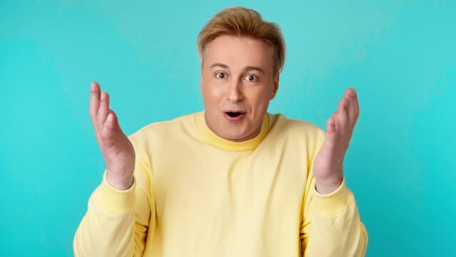 Surprised man with amazed wow expression. Excited guy with open mouth pleasant surprise emotion in yellow sweatshirt looking at camera, oh my god. Indoor studio shot isolated on blue background.