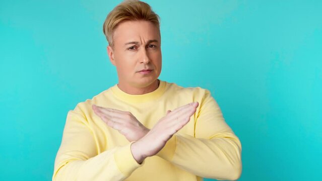 Displeased man says no in stop gesture crossing hands. Guy doing negative sign nodding no, angry face, rejection expression in yellow sweatshirt. Indoor studio shot isolated on blue background.