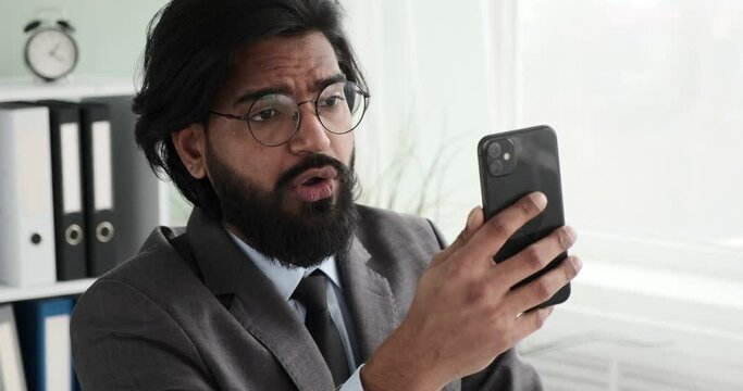 An Indian businessman in glasses sitting at his desk, intently communicating via video call on his phone. He appears focused in the conversation, discussing important business matters