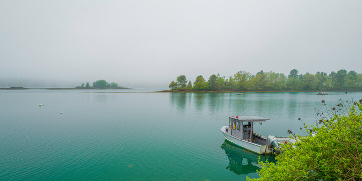 The moored trawler, trees on islands, and water reflections on the calm seawater. Tranquil Maine coastal road seascape over Casco Bay on Bailey Island on a foggy morning.