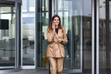 Young successful confident woman entrepreneur, CEO or an office worker talking on mobile phone leaving the business center, looking away and smiling