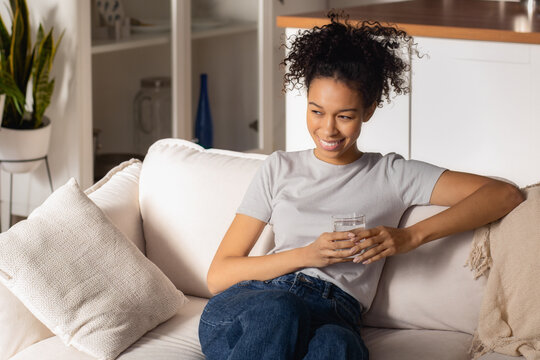 Young beautiful African American woman holding a glass of clean fresh water sitting on the sofa, relaxation at home, looking away and smiles