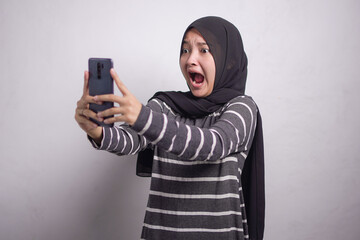 Shocked young asian woman in black and white sweater and hijab using on mobile phone and holding mouth with hand isolated over white background