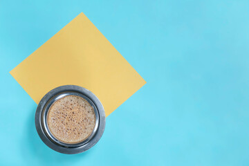 Cup of filter coffee with yellow note paper on blue background, top view