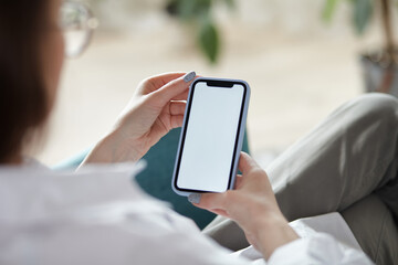 Mock up white screen blank mobile phone in woman hands holding sitting on armchair at home Back view