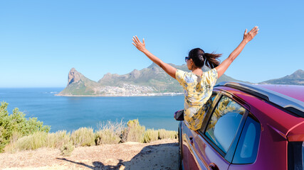 Obraz premium woman outside a car window with hands up, a car at Chapmans Peak Drive in Cape Town South Africa looking out over the ocean. women on a road trip garden route South Africa with renal car