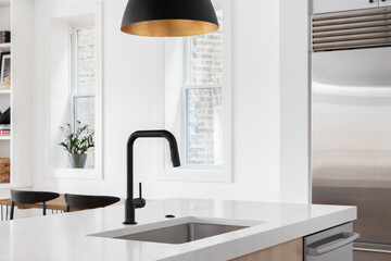 A kitchen sink detail with a black faucet under a black and gold pendant light, white oak island, white countertop, and decorations in the background.