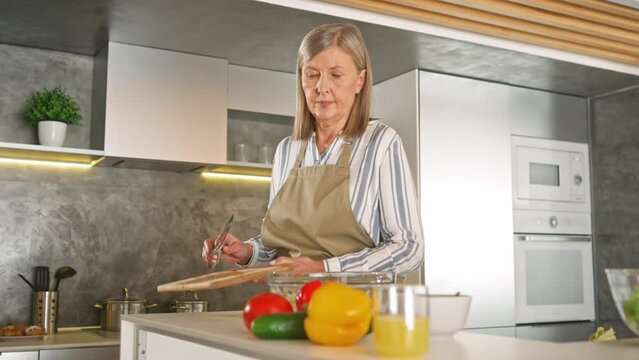 Pretty Caucasian woman with gray hair preparing salad for her lovely husband or kids. Charming female with apron chopping ripe cucumber and putting in plate with fresh vegetables. Cooking proccess.