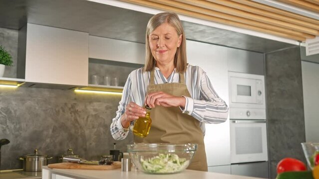 Good-looking mature woman with apron cooking fresh salad in cozy kitchen. Adding olive oil or sause to ripe vegetables. Preparing delicious dishes for family. Healthy nutrition concept.