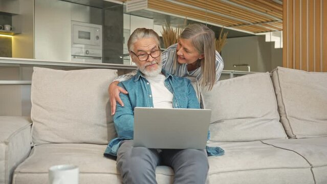 Mature Caucasian man using laptop while sitting on couch in living room. Charming wife coming up behind husband and hugging him. Showing tenderness and love to her partner. Relationship concept.